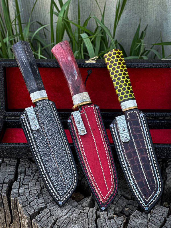 Три ножа и три кожаных ножна рядом на древесной поверхности: разные цвета рукоятей и аккуратная строчка на ножнах.