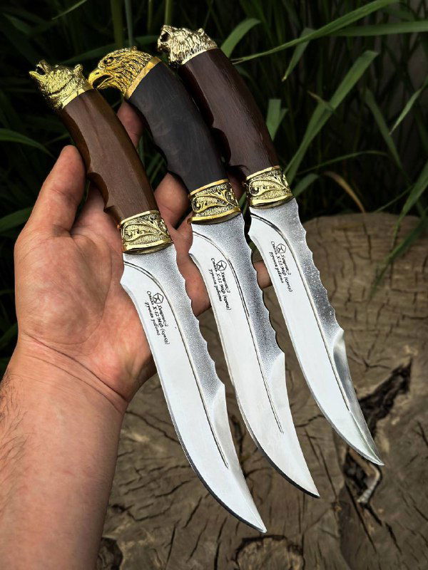 Три ножа в руке демонстрируют форму клинка и рукояти из граба и ореха с декоративной латунной обкладкой на фоне пней и травы.