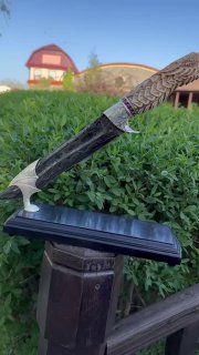 Авторский нож под углом: видно клинок с узором и серебряную гарду, рукоять из рога лося вырезана в форме дракона, фон с домом и кустами.