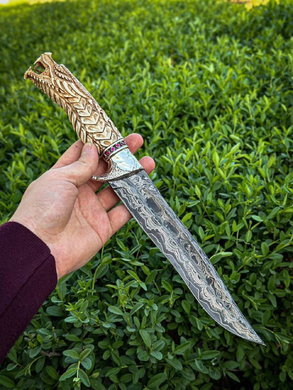 Рукоять ножа крупным планом: резьба по рогу лося в образе дракона, вставки фианитов в глазах и акценты окраса, фон зелёная листва.