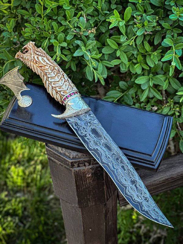 Клинок авторского ножа с выраженным узором и изгибом на подставке, видно соединение гарды и рукояти, фото на зелёном фоне.