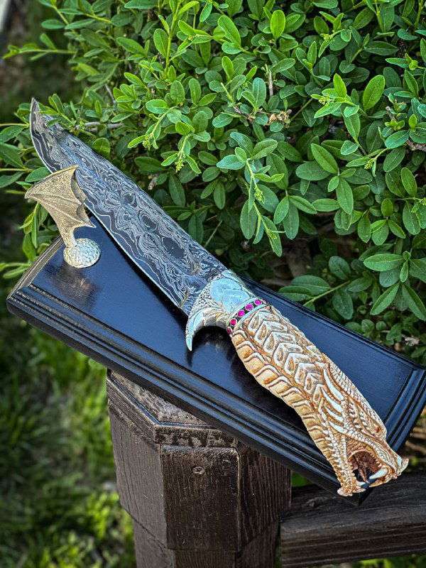 Нож «Дракон» лежит на подставке: полный вид клинка с узором и резной рукояти из рога лося, фон — кусты и садовая обстановка.