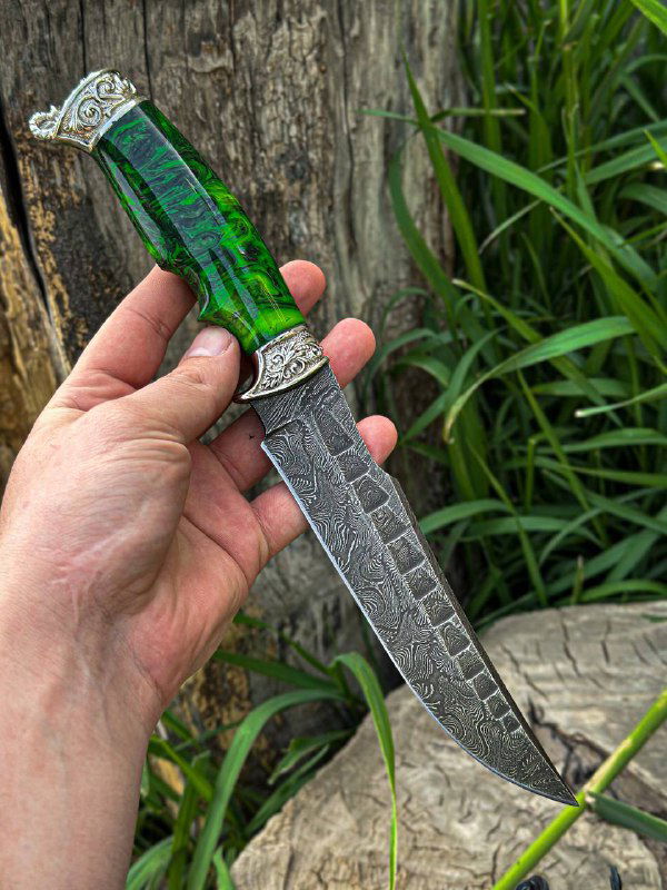 Рукоприкладное фото: нож «Скорпион» с зелёной акриловой рукоятью в руке мастера, видно рисунок дамасской стали на клинке и узоры на гарде.