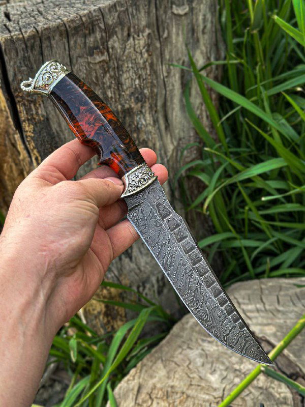 Рукоприкладное фото ножа с красно-чёрной акриловой рукоятью на фоне травы и древесины, показана длина и форма клинка модели.