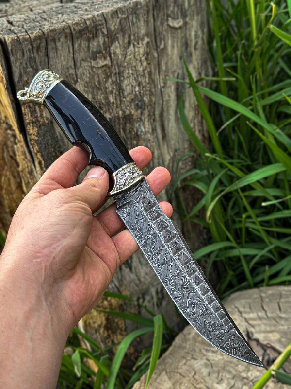 Фотография ножа с чёрной акриловой рукоятью: дамасская текстура клинка четко видна, фон — коряга и трава, демонстрация фактуры металла.