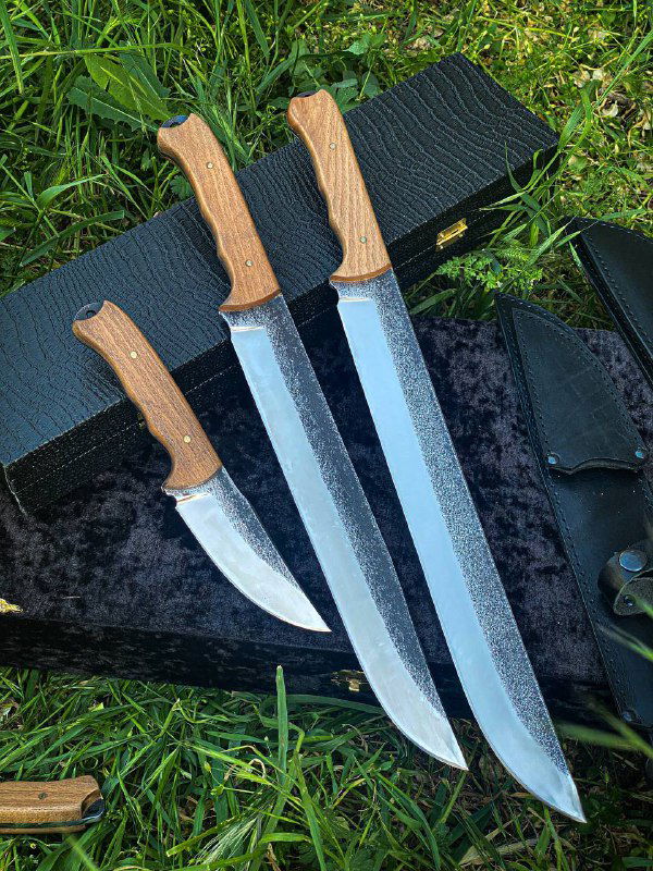 Три охотничьих ножа разного размера лежат рядом на траве: видны фактура клинков из Х12, одинаковые деревянные рукояти и кожаные ножны.
