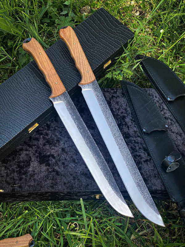 Два длинных клинка из стали Х12 с ореховыми рукоятями рядом на чёрной подложке и траве, видны спуски и текстура металла.