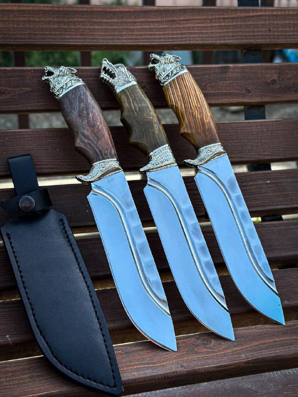 Три ножа на деревянной скамье в комплекте с кожаной ножной: видна древесная текстура рукоятей, декоративные мельхиоровые детали и форма клинка с профилем для охоты/туризма