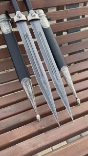 Крупный план трёх дамасковых клинков, видно рисунок стали и сходное оформление рукоятей из черного граба и мельхиора, расположение на деревянной скамье