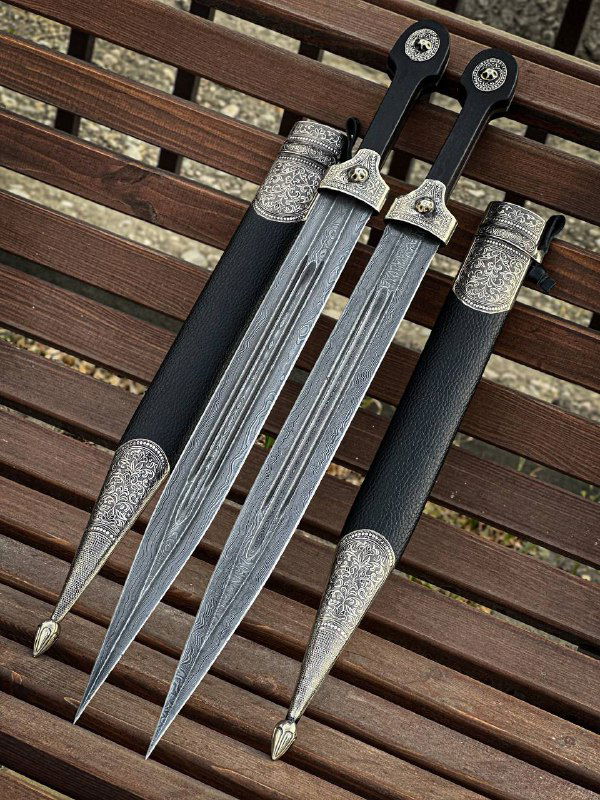 Фронтальный ракурс боевых кинжалов: видны лезвия с узором дамаска, орнамент мельхиоровых накладок и кожаные ножны на фоне деревянной поверхности