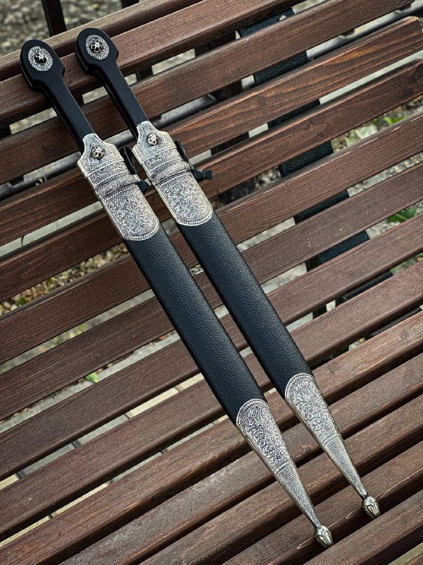 Общий вид двух кинжалов в ножнах с декоративными навершиями и текстурой кожаных ножен на фоне древесины, демонстрация полного комплекта