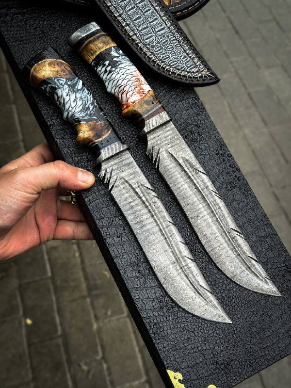 Пара ножей «Перо» с дамасковыми клинками, оформленные в кожаной подложке; видны сложный рисунок дамаска и симметричные формы клинков, подготовка к упаковке.
