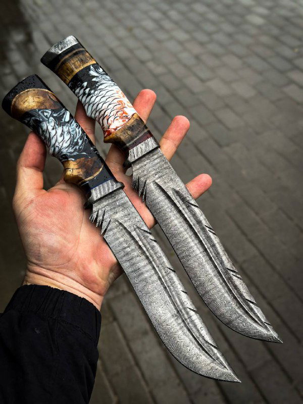 Две руки держат парные дамасковые ножи «Перо»: видно текстуру клинков, переходы металла и общую длину изделий, фон — влажная брусчатка.