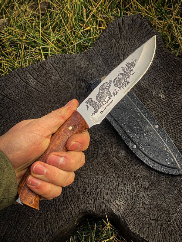 Крупный план руки, сжимающей рукоять ножа из кавказского ореха; на заднем плане кожаные ножны и трава.