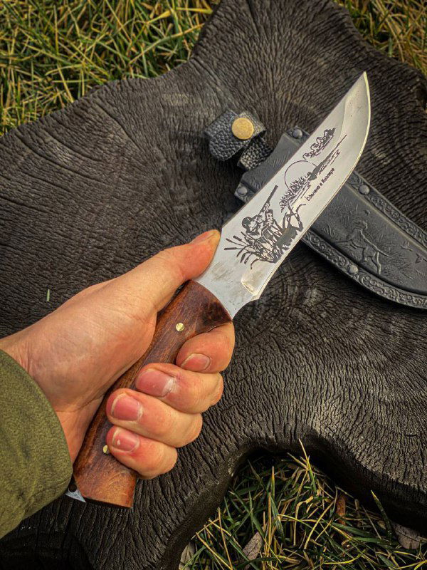 Детальное фото режущей части ножа с рисунком на лезвии и частично видной рукоятью из дерева на фоне шкуры.