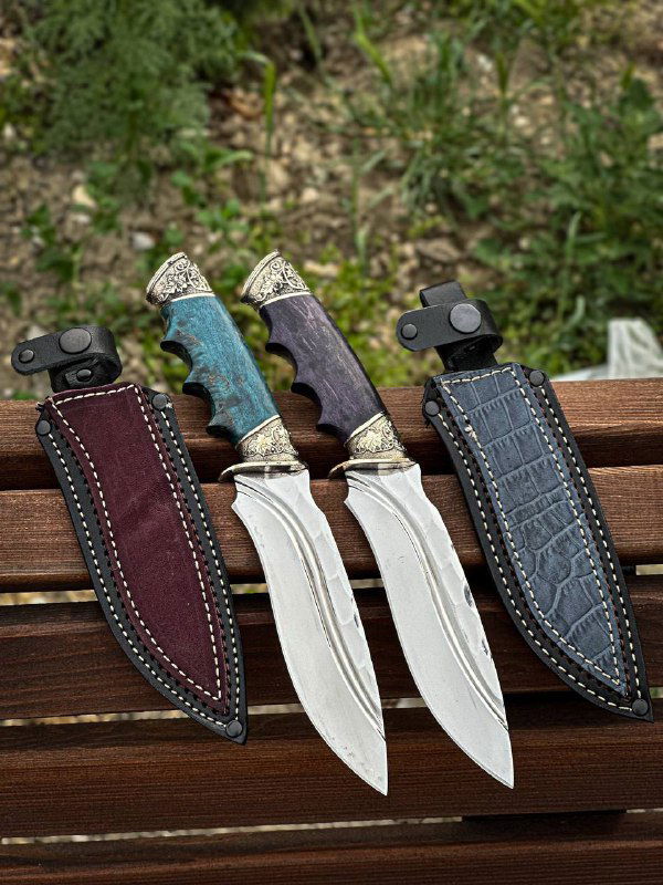 Три ножа рядом с кожаными ножнами на деревянной поверхности: видно боковой профиль клинков, швы ножен и отделку мельхиоровых гард.