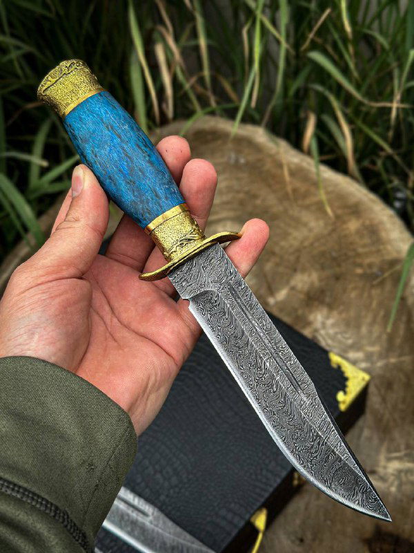 Крупный план ножа «Разведчик» в руке: дамасковая текстура клинка, латунная гарда и синяя стабилизированная карельская берёза на фоне травы и пенька