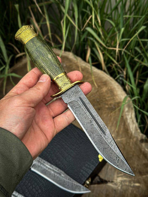 Рука держит нож «Разведчик» с зелёной стабилизированной карельской берёзой на рукояти, видна дамасковая структура клинка и латунная гарда на естественном фоне