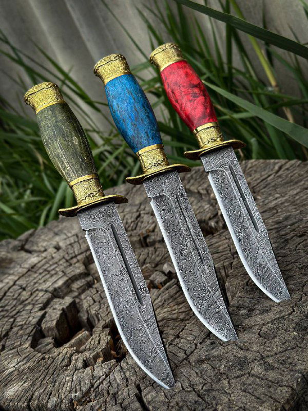 Три ножа «Разведчик» установлены вертикально на пеньке: видна различная текстура рукоятей, дамасковая поверхность клинков и латунные гарды на природном фоне