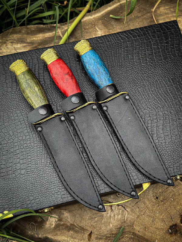 Три ножа «Разведчик» с одетыми кожаными ножнами, рукояти красного, синего и зелёного цветов видны сверху на тёмной поверхности, акцент на ножнах и фурнитуре