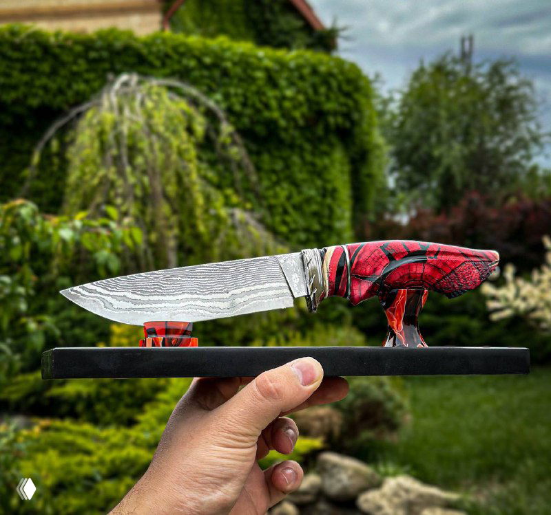 Ручной нож с ярко-красной стабилизированной рукоятью и дамасским клинком; макуме-элементы и монтажная скрутка видны на фоне зелёного ландшафта и камней.