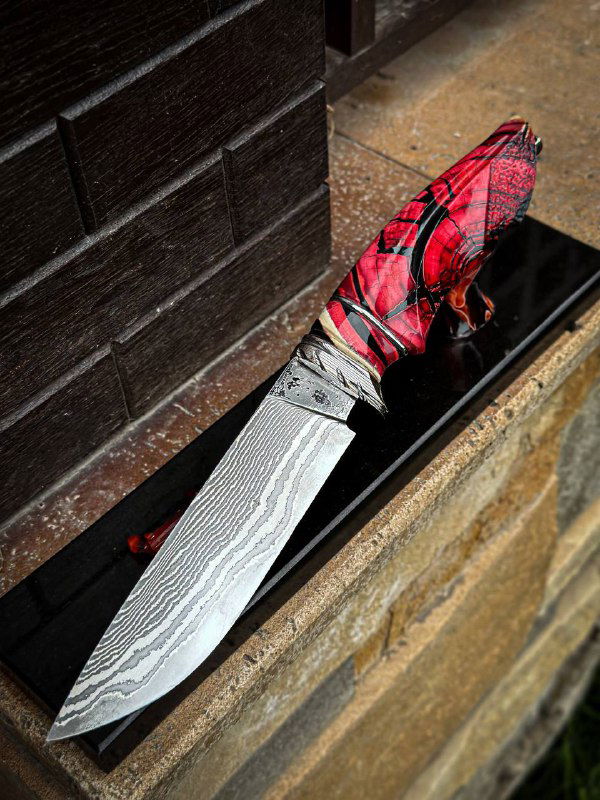 Крупный план авторского ножа с дамасским клинком и стабилизированной рукоятью из бивня мамонта; нож лежит в черном лотке на каменной поверхности.