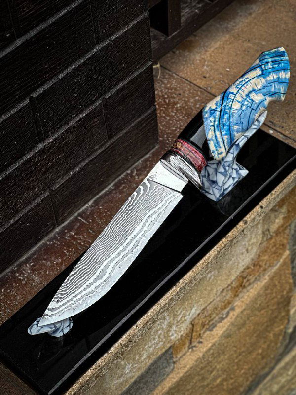 Авторский нож с изогнутой рукоятью из стабилизированного бивня мамонта и художественной гардой из макуме, установлен на чёрной подставке у стены мастера.