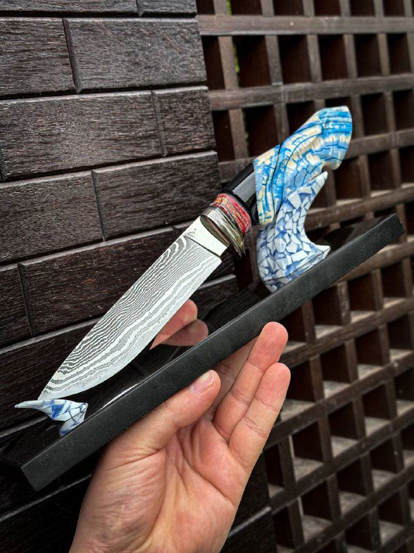 Нож мастера: дамасская клинковая текстура, рукоять из бивня мамонта с декоративными вставками, показан на гранитной подставке в руке владельца.