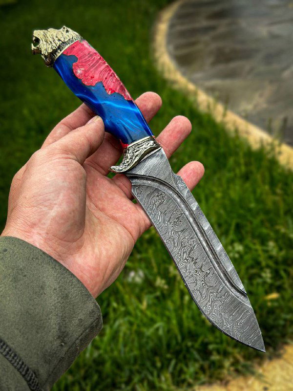 Крупный план одного ножа в руке: дамасковый клинок с характерным рисунком и синяя рукоять из карельской берёзы с мельхиоровым литьём у обуха.