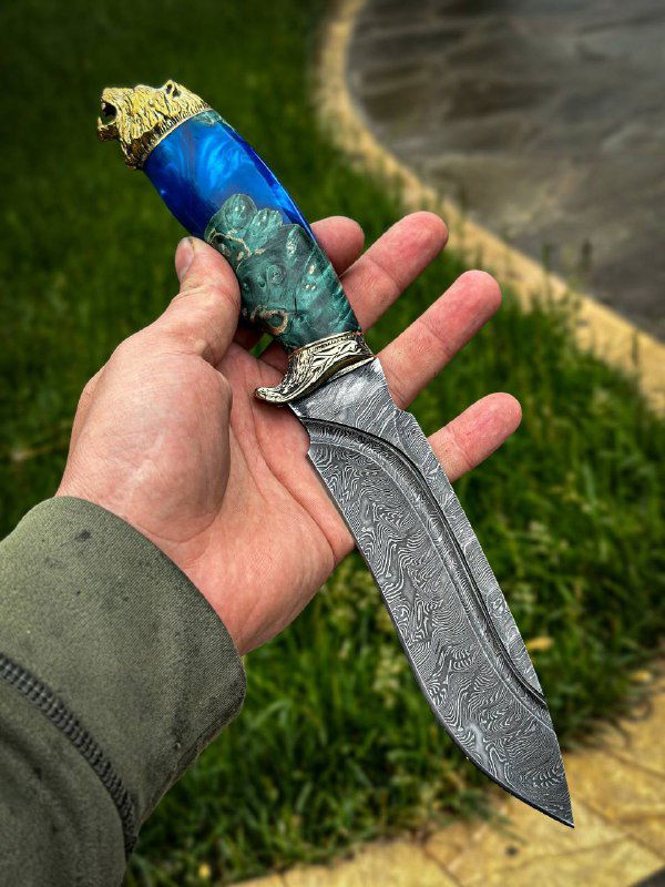 Ещё один ракурс ножа в руке: видно детализированное мельхиоровое литьё, переход от клинка к рукояти и зелёно-голубые оттенки вставки на рукояти.