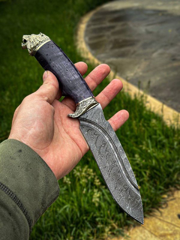 Чёрная рукоять ножа в руке на фоне зелёной травы и воды: видно текстуру карельской берёзы, контраст с дамасковым клинком и мельхиором у гарды.