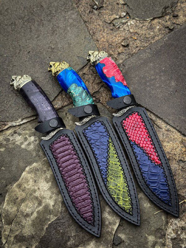 Три ножны из кожи с цветными вставками лежат рядом с ножами: видны тиснение и вставки, подходящие под цвета рукоятей, комплектный вид для продажи.