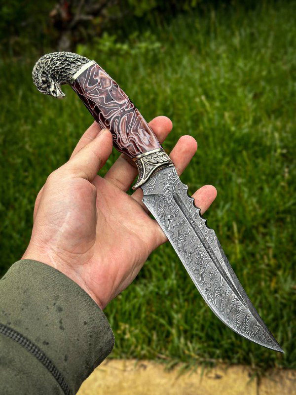 Нож «Орлан» в руке мастера с розовато-мраморной акриловой рукоятью, видно оформление гард и рисунок дамаска по лезвию на природе.