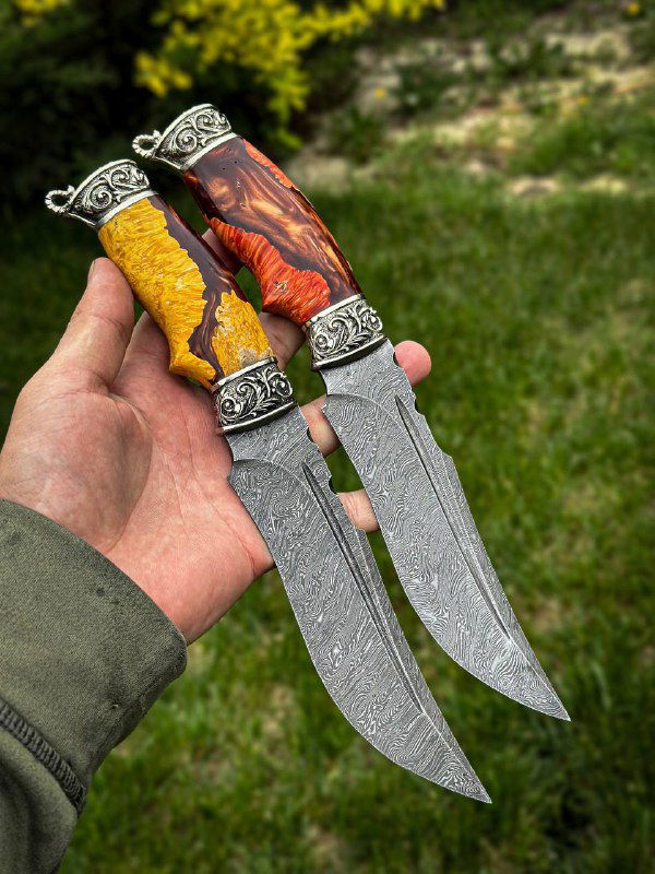 Крупный план двух ножей «Скорпион» в руке на зелёном фоне; видно узор дамаска на лезвии, оформление фурнитуры и древесную текстуру карельской берёзы рукоятей.