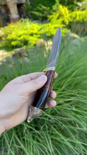 Боковой вид ножа «Волк» в руках: профиль клинка и аккуратная посадка гарды, рукоять из тёмного акрила, фон — летний сад и зелень.