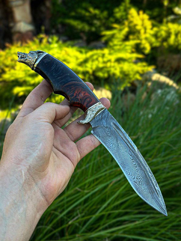 Ещё один ракурс ножа «Волк»: отчётливый дамасский рисунок на лезвии, декоративная мельхиоровая гарда и тёмная акриловая рукоять, держится в руке над травой.