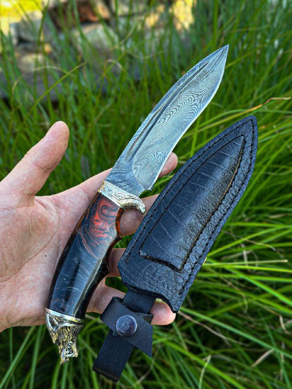 Нож «Волк» в ножнах: клинок с дамасским узором частично вынут, видны кожаные ножны чёрного цвета и отделка гарды, фон — зелёная трава и сад.