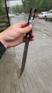 Вид ножа в руке: тонкий дамасковый клинок, декоративная рукоять с акриловой шишкой и металлические детали, на фоне городской тротуар.