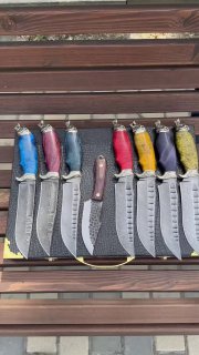 Групповая фотография ножей «Волк» на деревянной поверхности с видимыми текстурами клинков и пятнами цветных рукоятей, один нож в центр.