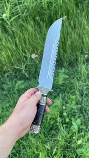 Крупный план одного ножа: гладкий полированный клинок, пила на обухе, художественные мельхиоровые детали и стабилизированная чёрная рукоять, фон — размытая зелёная растительность.