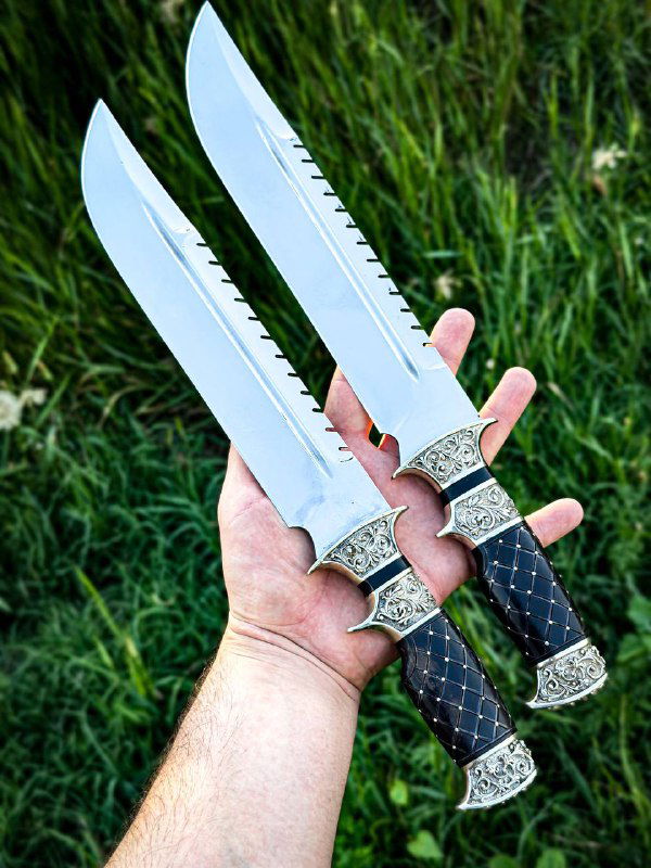 Два ножа рядом в одной руке: одинаковая форма клинков с зубцами на обухе, идентичные чёрные рукояти с узорным мельхиоровым оформлением, фон — летняя трава.