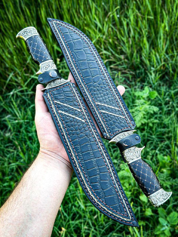 Авторские кожаные ножны тёмного цвета с выделанной фактурой и строчкой, рядом часть рукояти ножа с мельхиоровыми накладками, фотография на зелёном фоне.