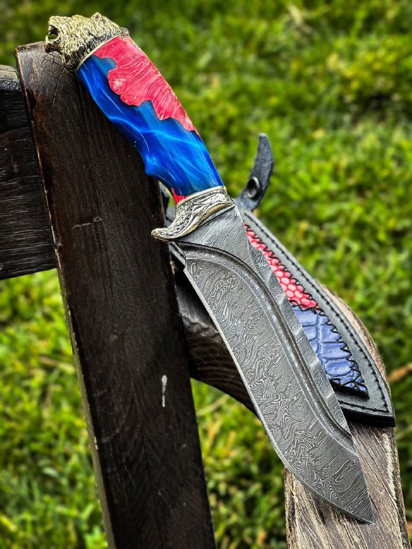 Крупный план одного ножа: дамасковое лезвие с выразительной текстурой, рукоять с красно-синими декоративными вставками и мельхиоровым навершием на зелёном фоне.