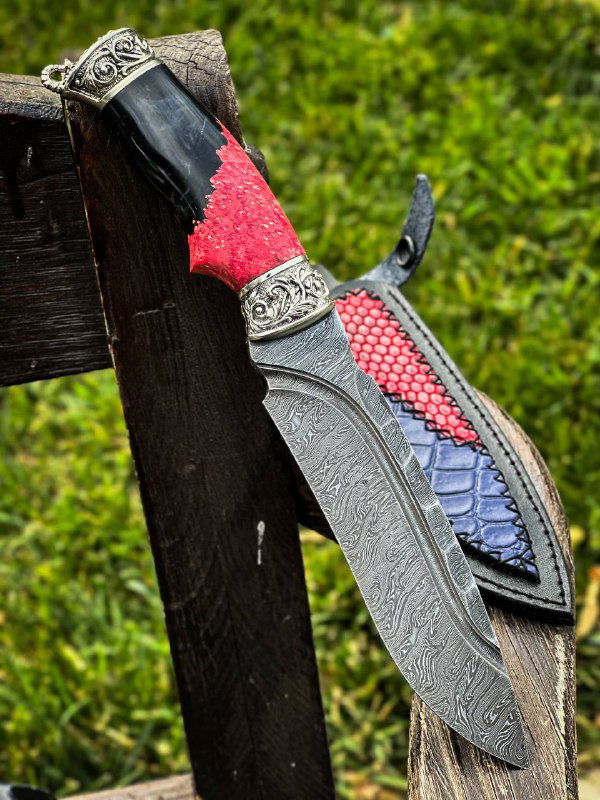 Ещё один крупный план ножа в ножнах: видно мельхиоровое литьё на гарде, красные вставки на рукояти и текстуру дамаскового клинка на фоне травы.