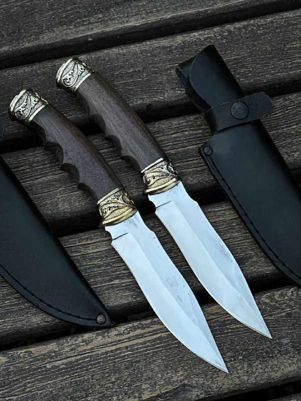 Два ножа «Жало» с рукоятями из стабилизированного ореха и мельхиоровыми гардами, рядом кожаные ножны на деревянной лавке.