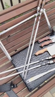 Длинные шампура и рукояти ножей, разложенные на лавочке рядом с мангалом, видна стальная поверхность шампуров и деревянные ручки.