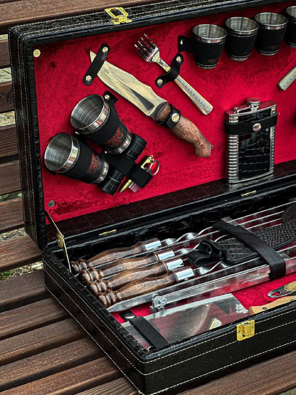 Крупный план наборов стаканов, ножей и фляжки на красной подкладке в раскрытом чемодане-наборе для шашлыка.