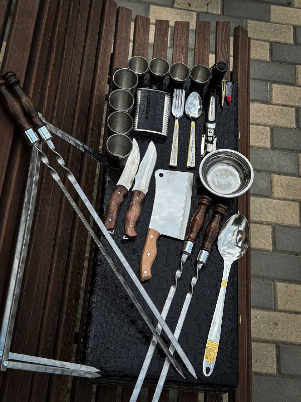 Разложенные на тёмной поверхности элементы набора: шампура, ножи, разделочная доска, столовые приборы и мелкие аксессуары.