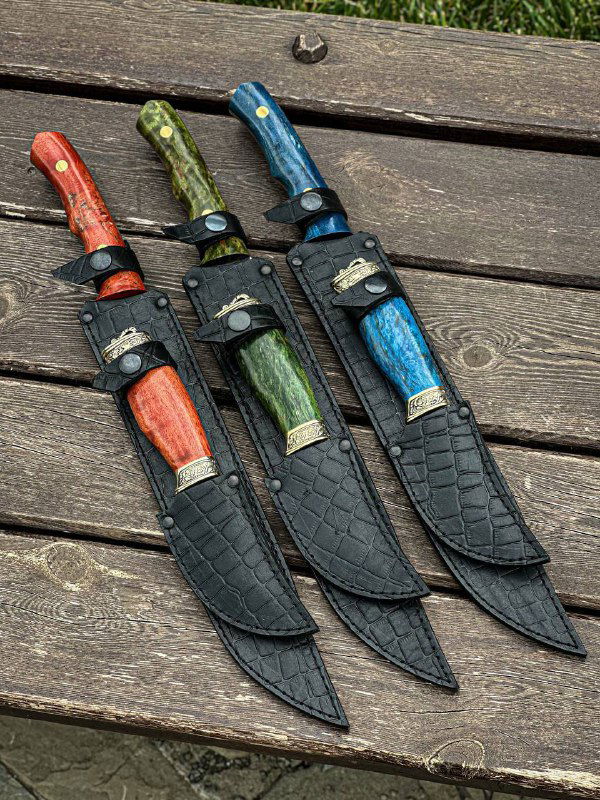 Четыре цельнометаллических ножа разных размеров с кожаными ножнами и цветными рукоятями из стабилизированной карельской берёзы, разложенные на деревянной поверхности.