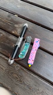 Вид сверху: три разложенных ножа на деревянной скамье — чёрно‑серый кинжал и два складных цветных ножа рядом друг с другом.